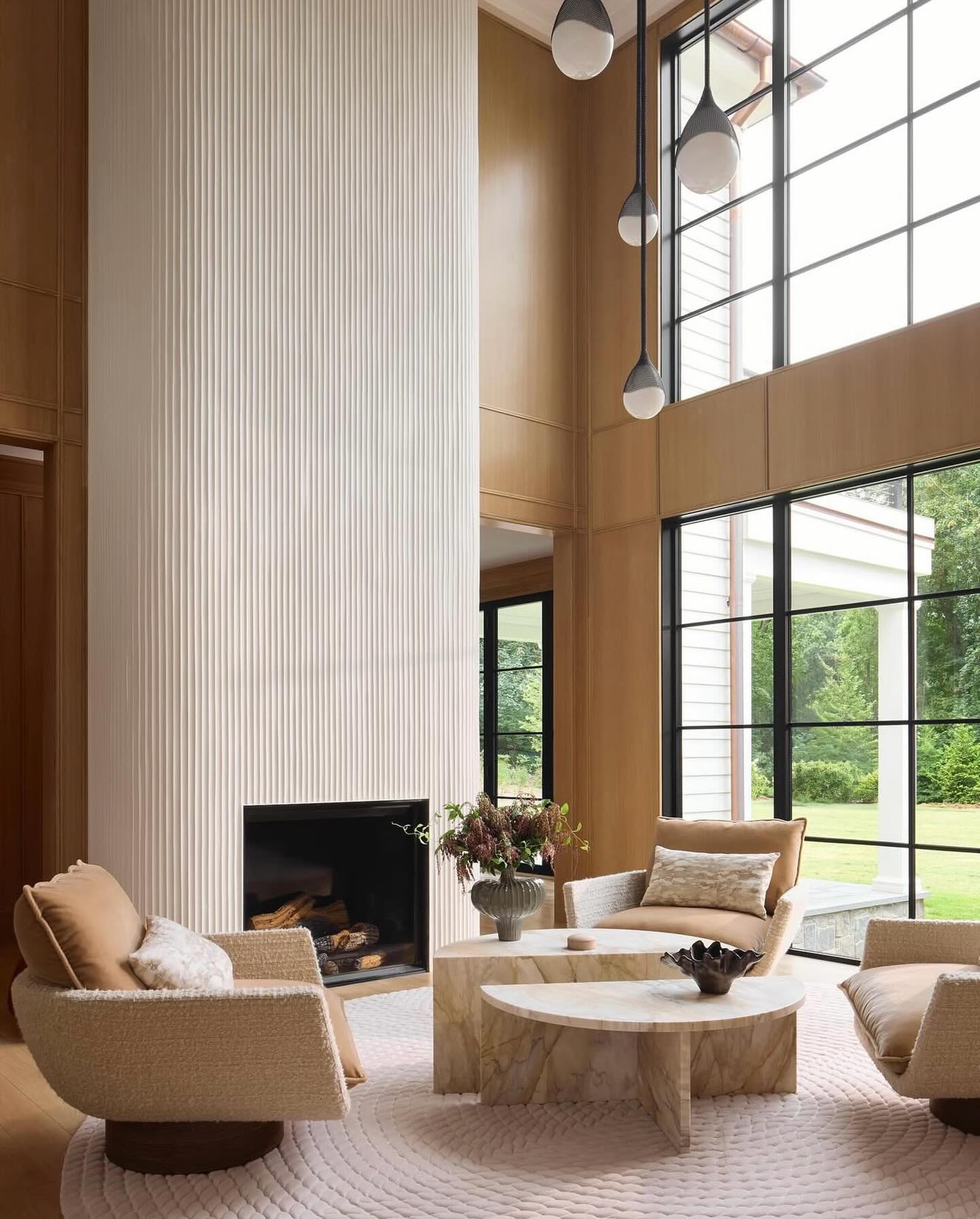 Contemporary modern farmhouse living room with double-height wood paneled walls, white fluted plaster fireplace, caramel leather and boucle swivel chairs, and half-moon marble coffee tables
