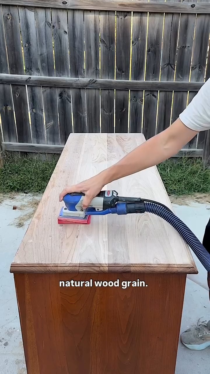 A blue square orbital sander with a dust extraction hose running across the dresser top, revealing pale raw wood grain, with a weathered wood fence in the background.