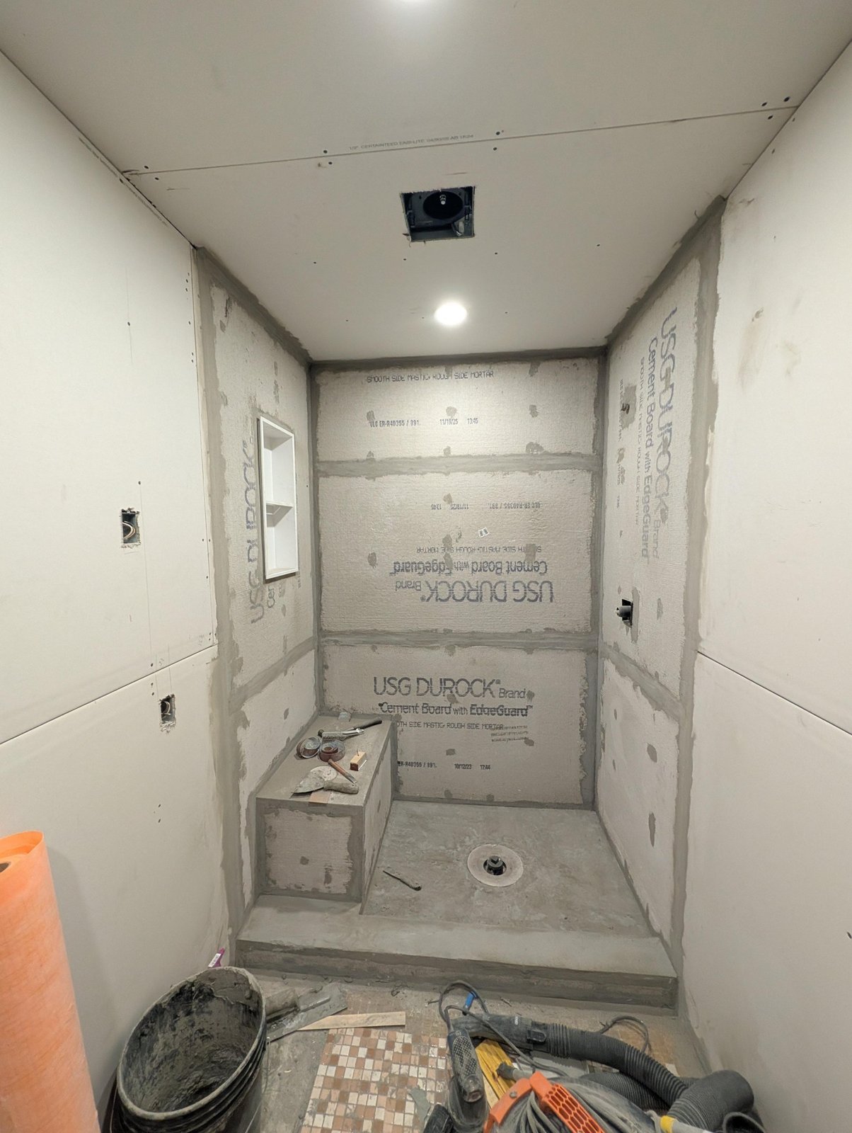 Completed Durock cement board walls inside the shower with taped and mudded seams, a built-in mortar bench, a recessed niche cut into one wall, and a shower drain in the center.