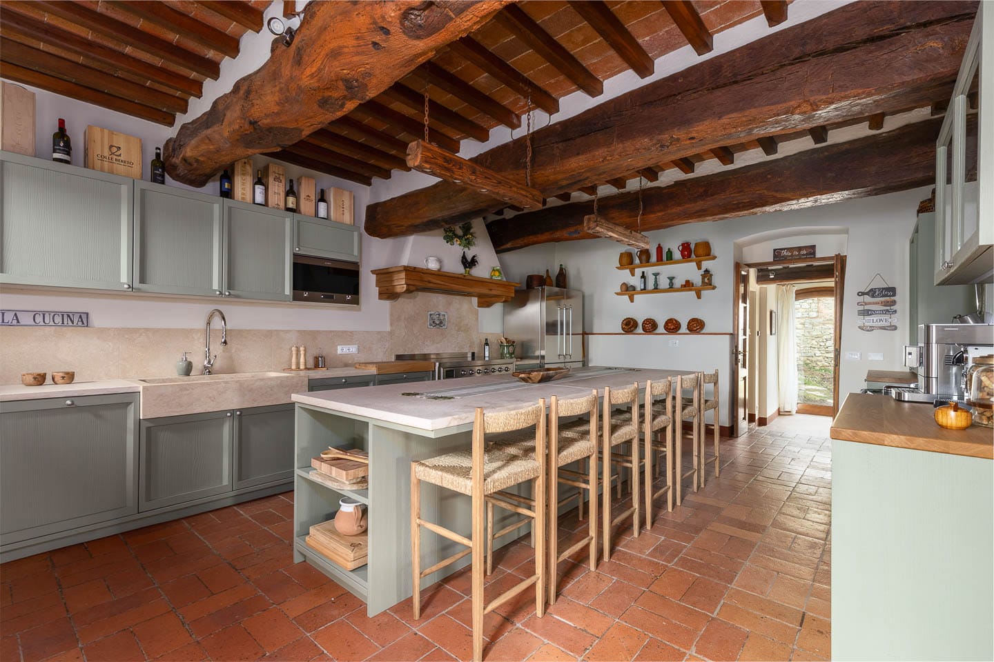 Authentic rustic Tuscan kitchen with original rough-hewn ceiling beams and terracotta tile ceiling, sage green glass-front cabinets, stone farmhouse sink, long sage island with rush-seat stools, and terracotta tile floor