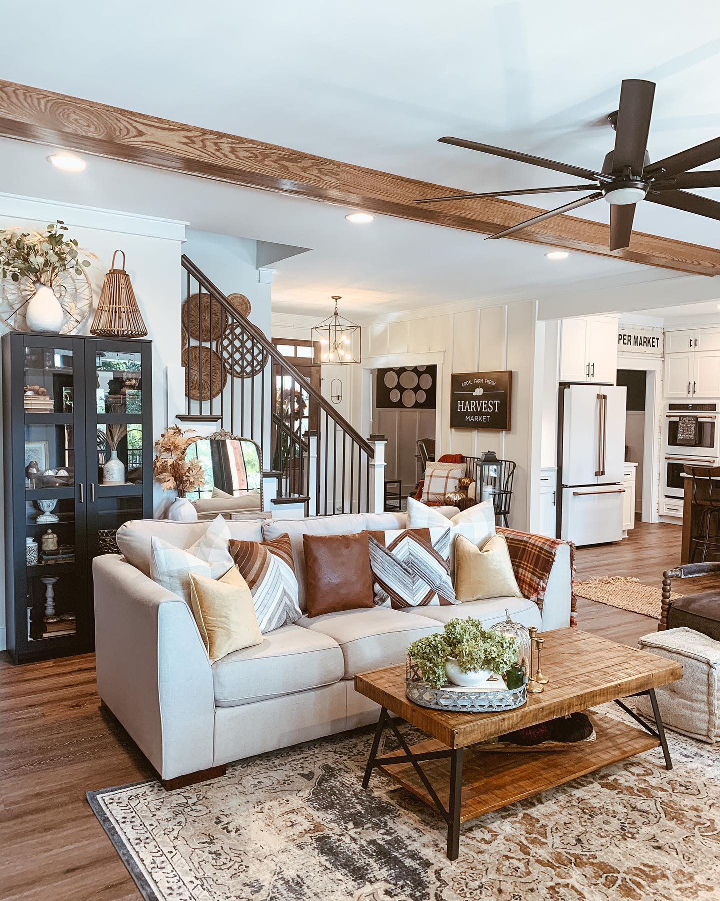 Modern farmhouse living room styled for fall with white sofa, cognac leather pillow, plaid pillow, mustard pillow, dark glass display cabinet, and vintage area rug under a wood beam ceiling