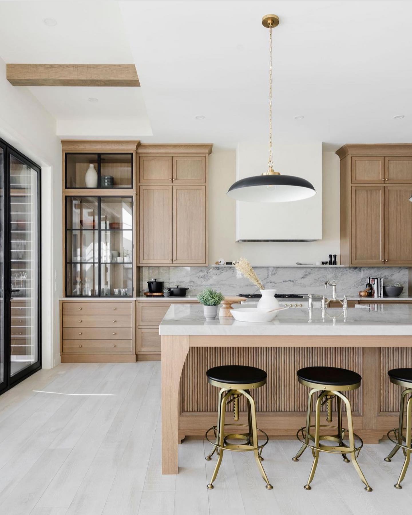 Light oak kitchen with a fluted vertical-grooved oak island, grey veined marble backsplash, large black and brass dome pendant, three brass bar stools with black leather seats, and a glass-front display cabinet