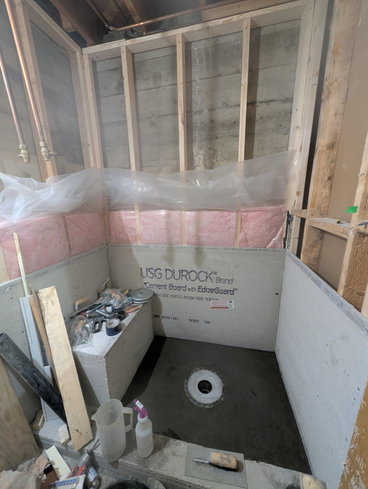Bathroom shower corner with fresh wood framing, pink insulation batts, Durock cement board going up on the walls, and a mortar shower pan with a center drain.