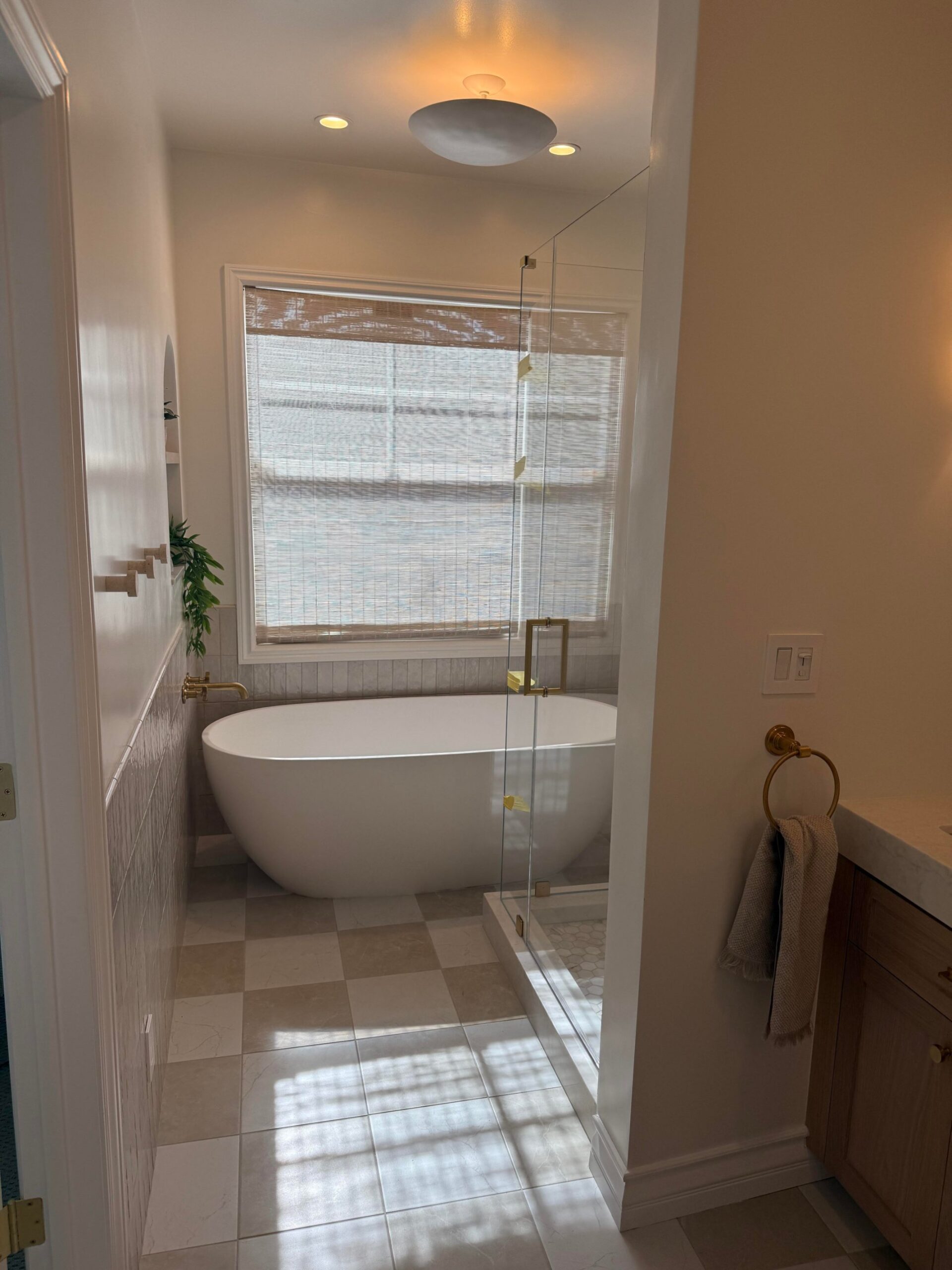 White freestanding soaker tub under a window with woven roller shade, cream and tan checkerboard marble floor, wall-mounted brass filler, and a glass walk-in shower to the right.