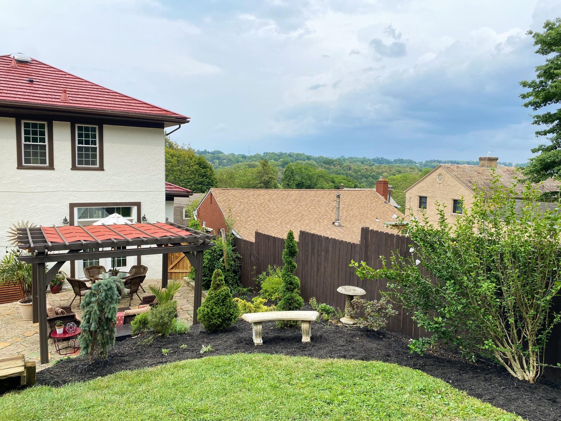 Wide view of the finished backyard with a pea gravel fire pit ringed by wooden Adirondack chairs and brick edging, a dark pergola with wicker lounge in the back, a dining spot under a cream umbrella, and string lights overhead.