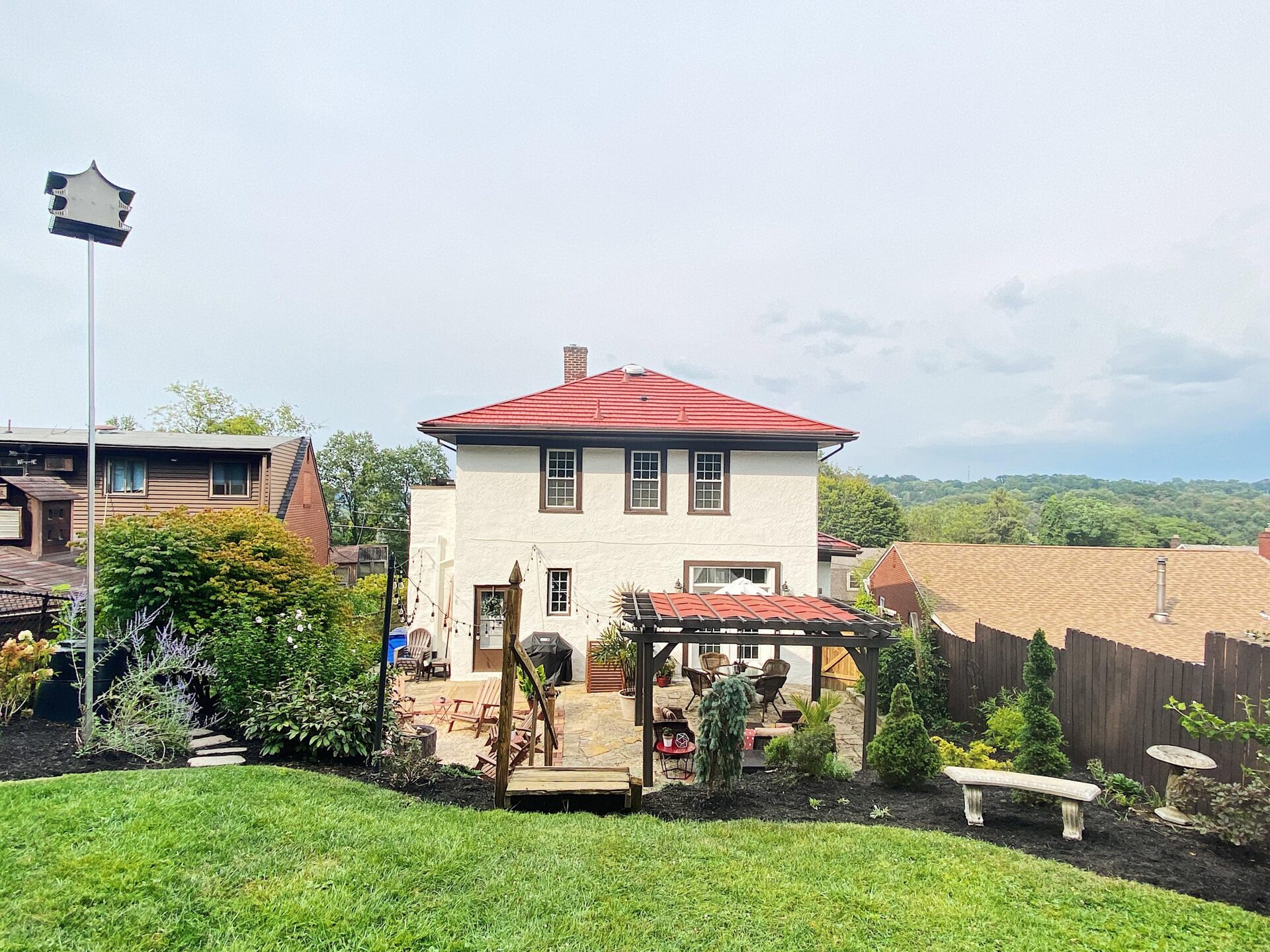 Stepping stone path through a mulched garden bed with purple Russian sage, white roses, and shrubs along the white stucco house, with Edison bulb string lights running from the house to a tall black garden pole and a wrought sun ornament on the stucco wall.