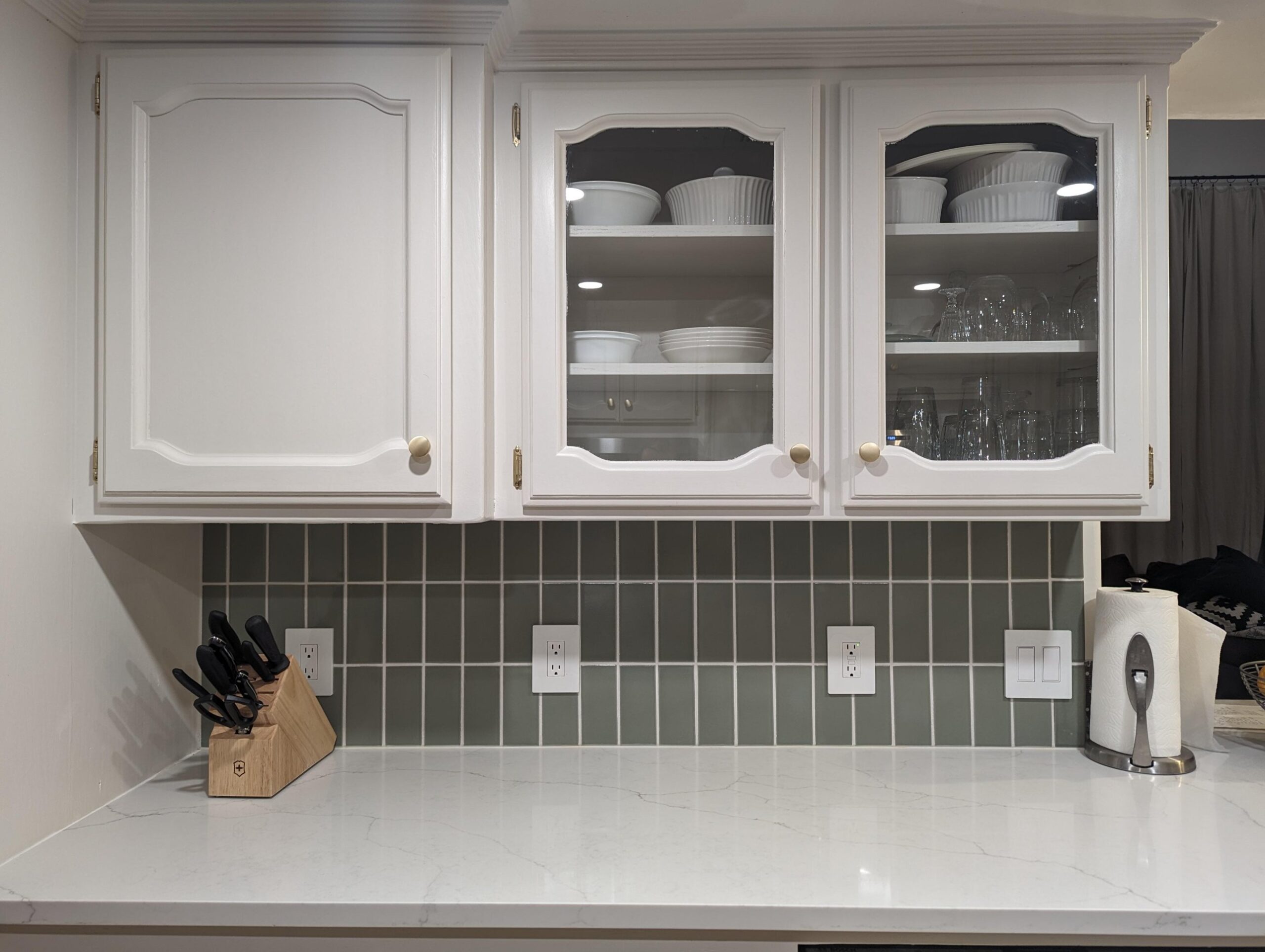 Close view of the glass-front upper cabinets painted white, stacked dishes and glassware visible inside, sage green vertical tile backsplash below with white outlet covers.