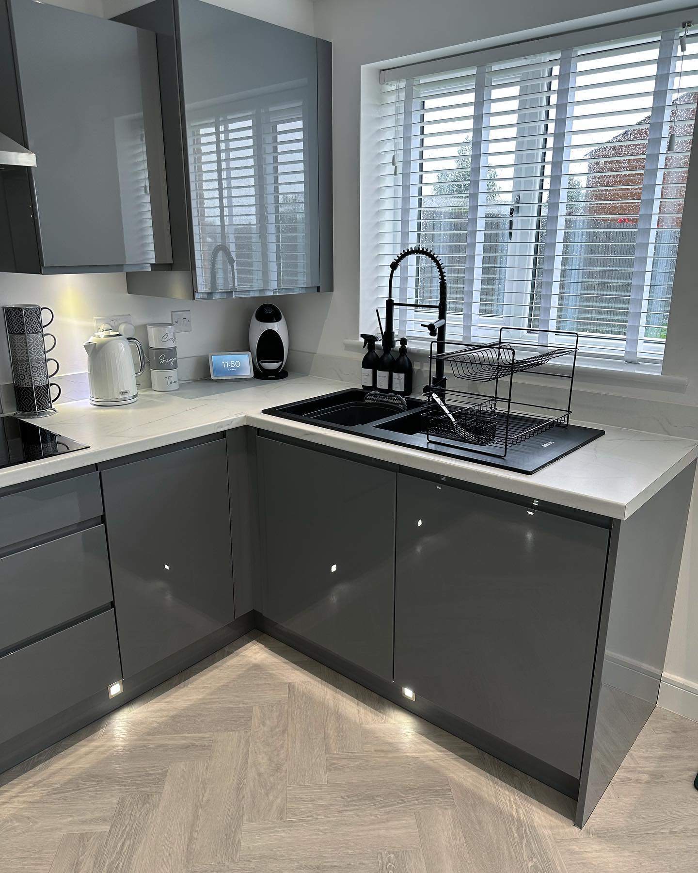 Mid-grey high-gloss handleless L-shape kitchen with white marble countertop, black double-bowl composite sink, black spring tap, white wood Venetian blinds, LED plinth lights under the cabinets, and grey herringbone wood-look flooring