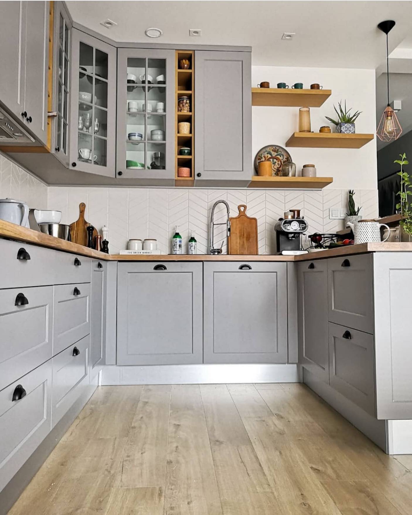 Light grey shaker kitchen with butcher block wood countertops, black cup pull handles, glass-front upper cabinets with brass trim, open wood floating shelves, chevron white tile backsplash, and natural oak flooring