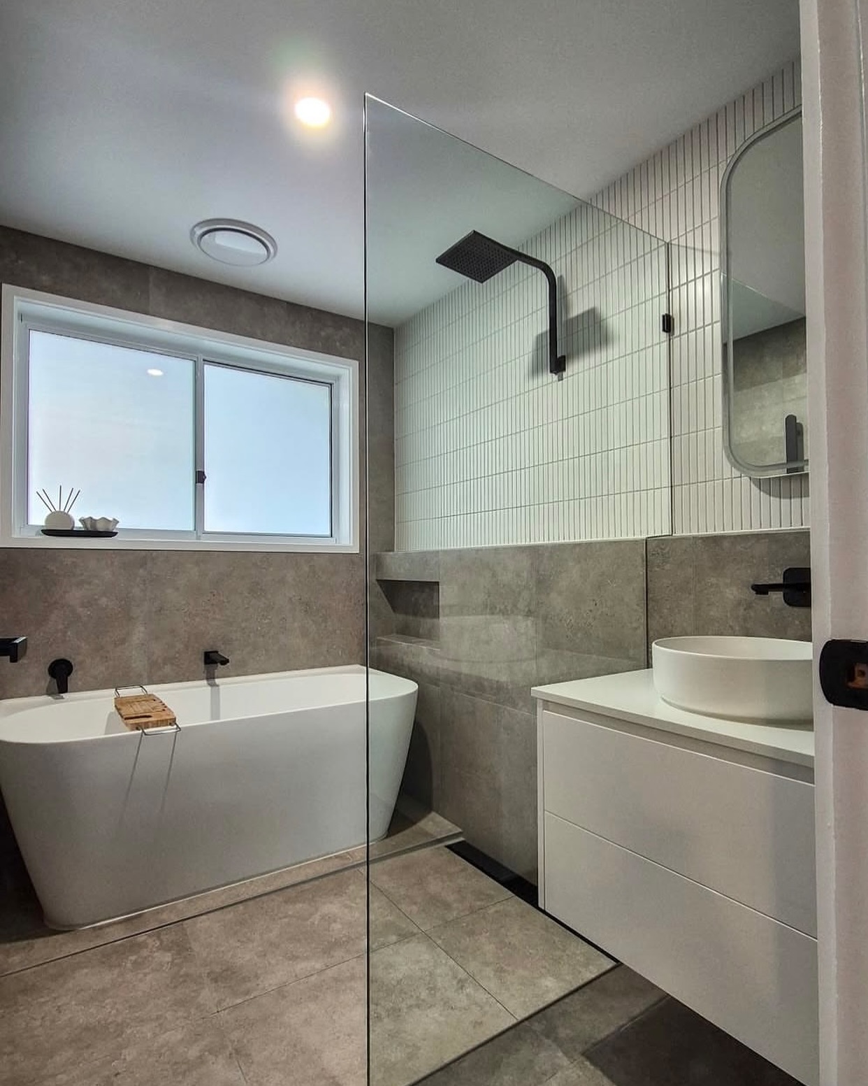 Small grey wet room bathroom with a freestanding white tub, vertical white kit-kat tile shower wall, black rain shower, floating white vanity with a round white vessel sink and a small corner window