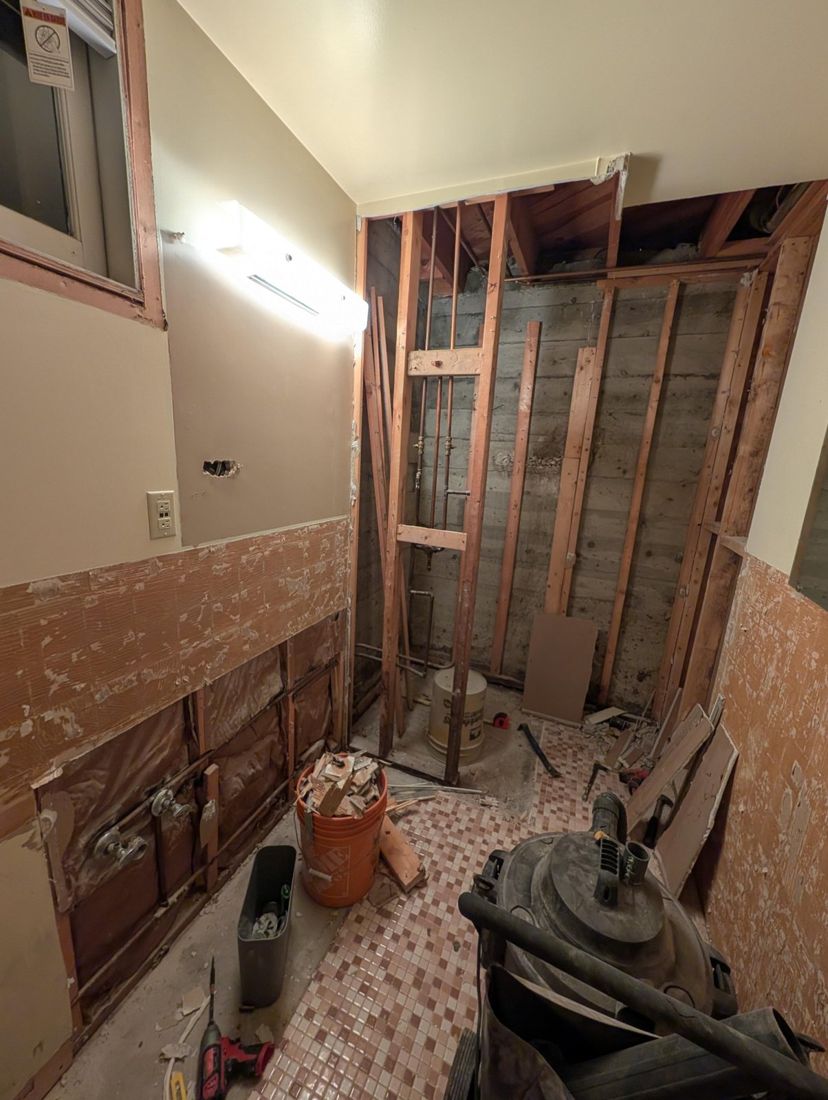 Bathroom mid-demo with mint-green wall tile ripped off, old pink mosaic floor still in place, exposed wood framing and copper pipes, buckets and tools on the floor.