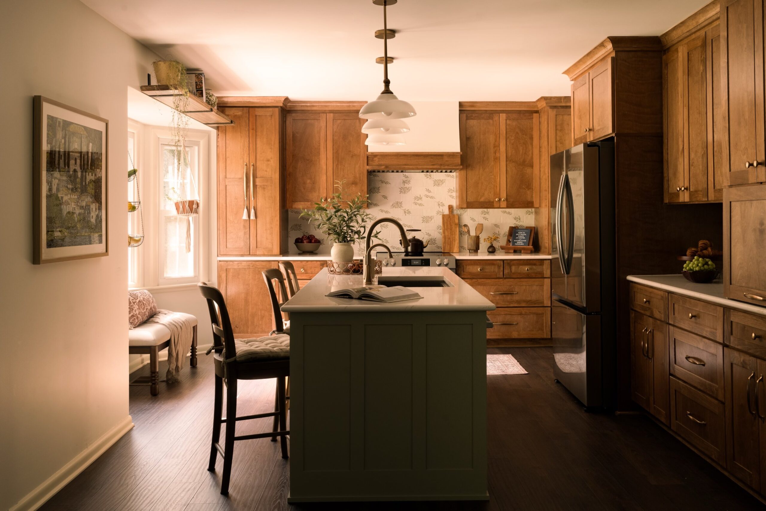 American Tuscan-style kitchen with honey oak cabinets, sage green painted island, patterned olive branch tile backsplash, white glass bell pendant lights, dark hardwood floors, and a window seat with Mediterranean art