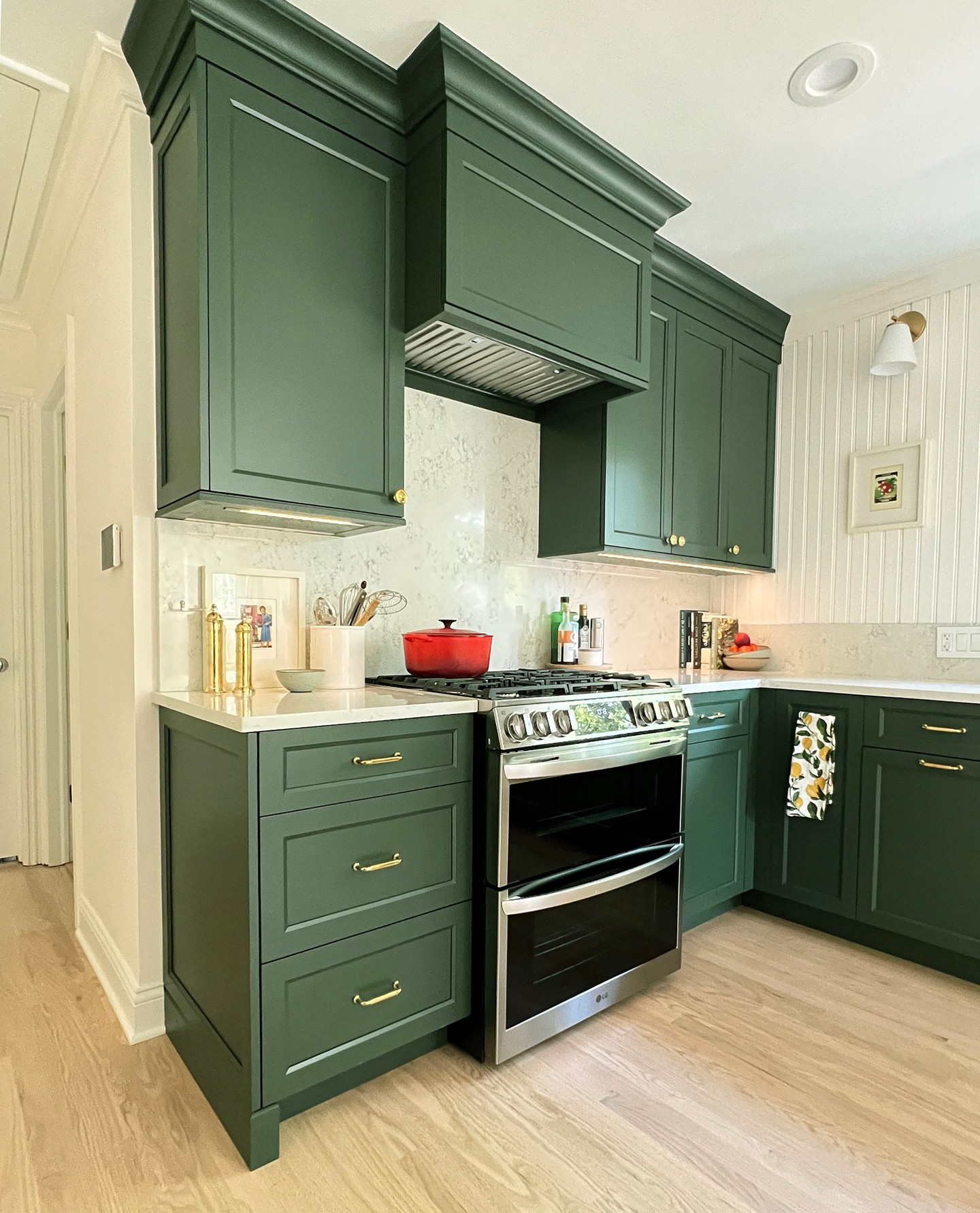 Hunter green shaker cabinets with brass bar pulls and knobs, a stainless steel LG double oven slide-in range, white quartz countertops with soft gray veining, a red Le Creuset dutch oven on the stove, a lemon-print tea towel on the oven handle, and a light oak floor