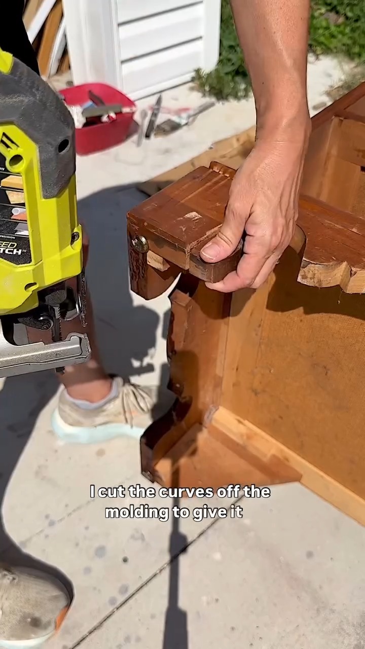 A yellow Ryobi jigsaw cutting through a curved wooden bracket piece at the base of the dresser, with the dresser tipped onto its back on a concrete surface and a hand steadying the piece being removed.