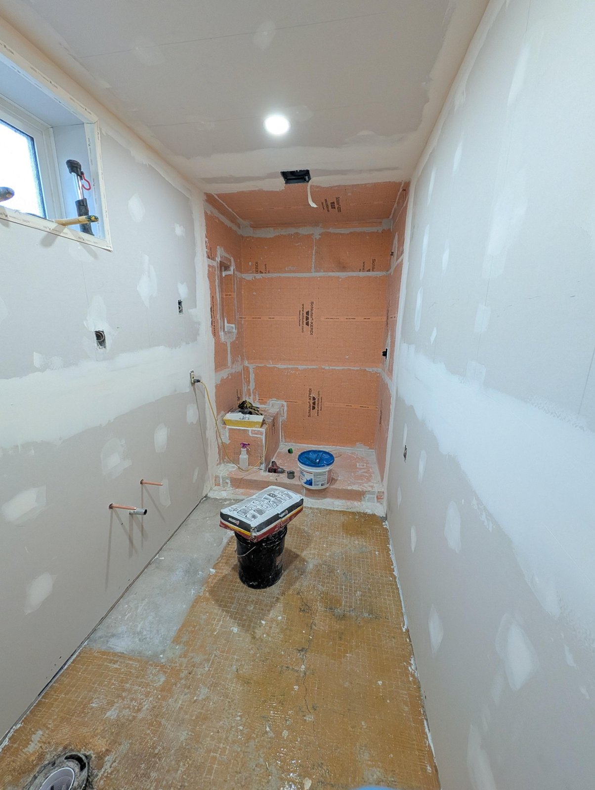 Shower walls and bench covered in orange Schluter Kerdi waterproofing membrane, with smooth drywall mudded around the shower opening and a bare concrete subfloor.