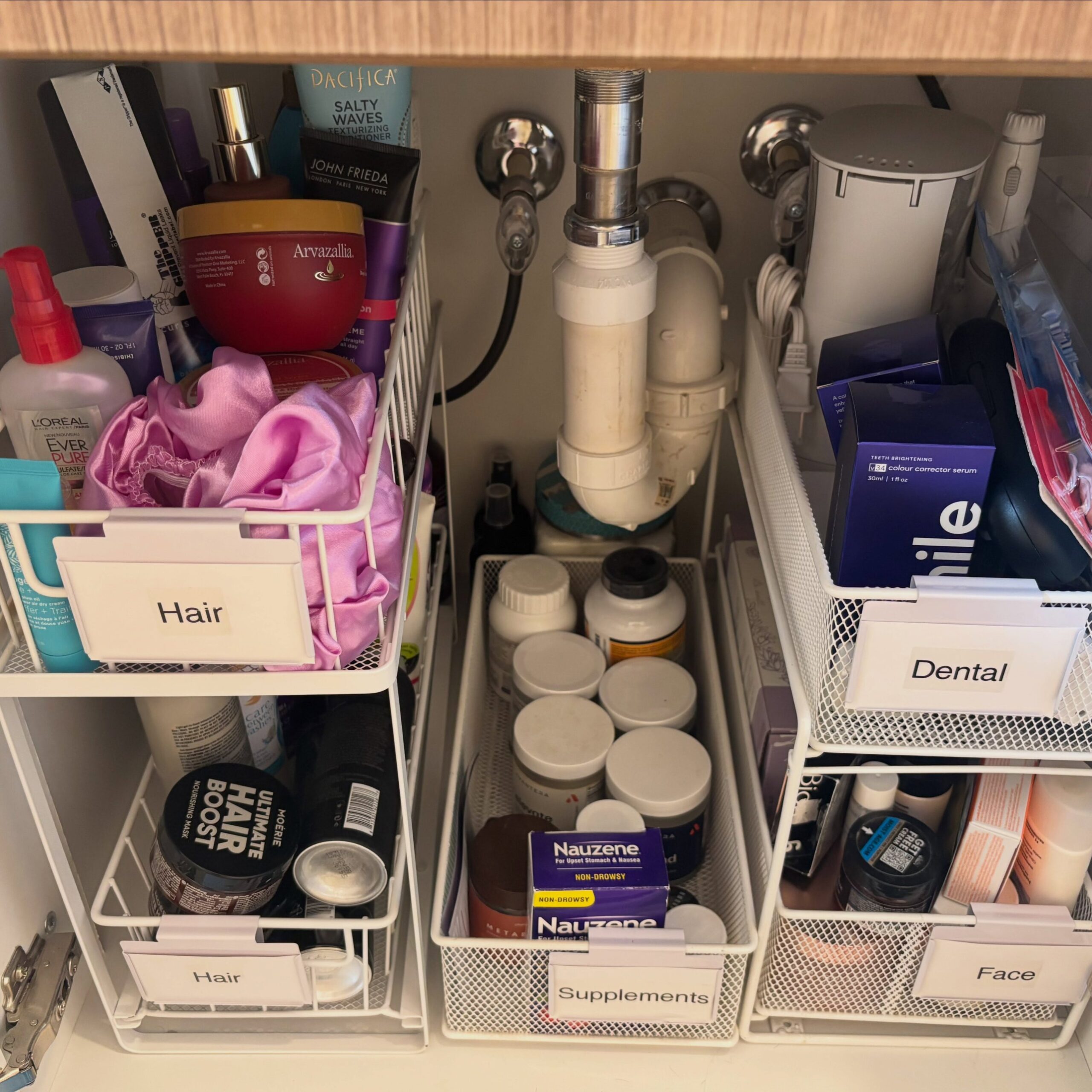 Three white wire two-tier sliding baskets under a bathroom sink, labeled Hair, Supplements, Dental and Face, holding hair masks, vitamin bottles, teeth whitening boxes and skincare