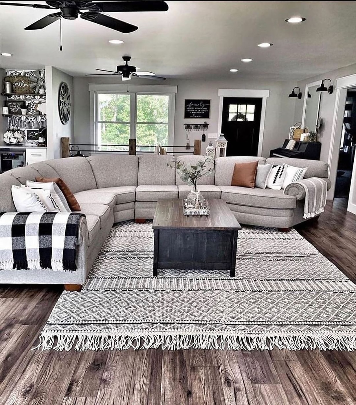 Spacious modern farmhouse living room with large curved gray sectional sofa, two black ceiling fans, patterned fringed rug, buffalo check throw, and dark wood coffee table