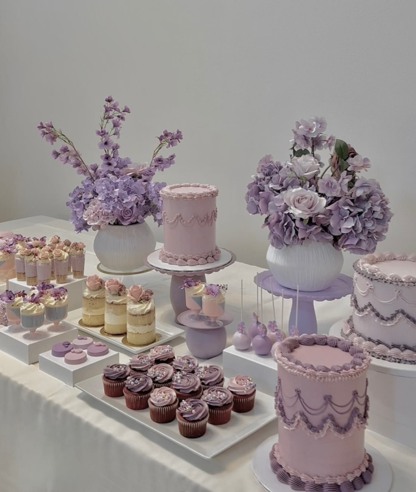 Lavender and pink vintage dessert table with ornate piped cakes, cupcakes in pleated liners, and purple hydrangea arrangements in white vases
