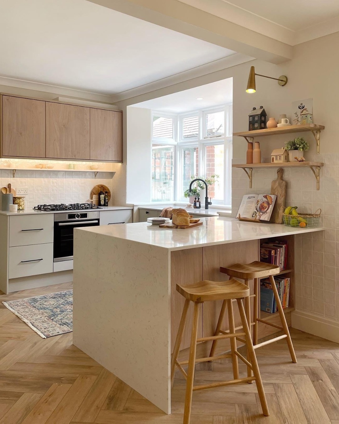 Cozy small kitchen with light oak upper cabinets, a white quartz-top peninsula with oak sides, cream square tile backsplash, open brass-bracket shelves, herringbone wood floors, and wooden bar stools