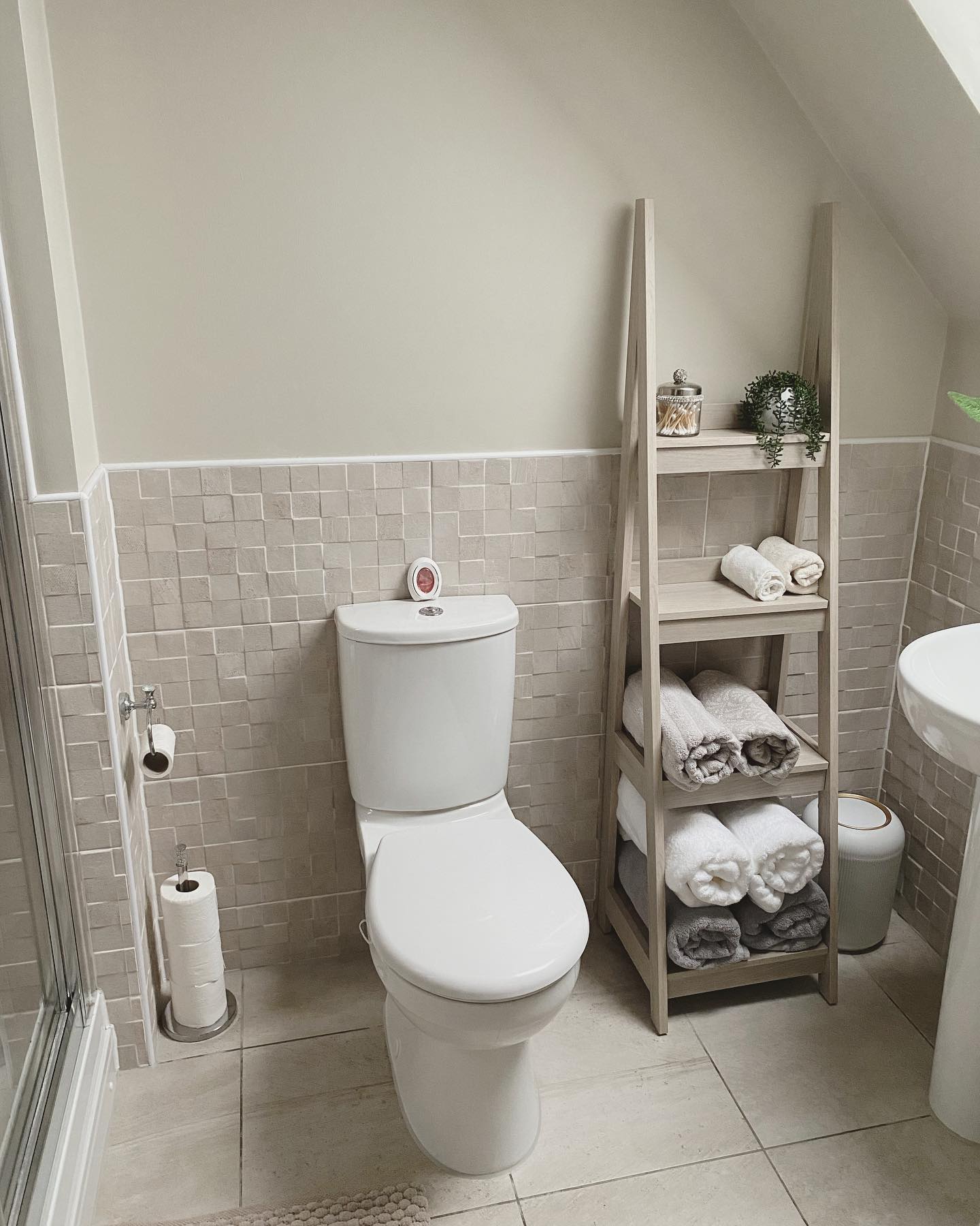 Light wood ladder tower shelf standing in the narrow spot between a toilet and sink in an attic bathroom with sloped ceiling and small stone tile wainscot