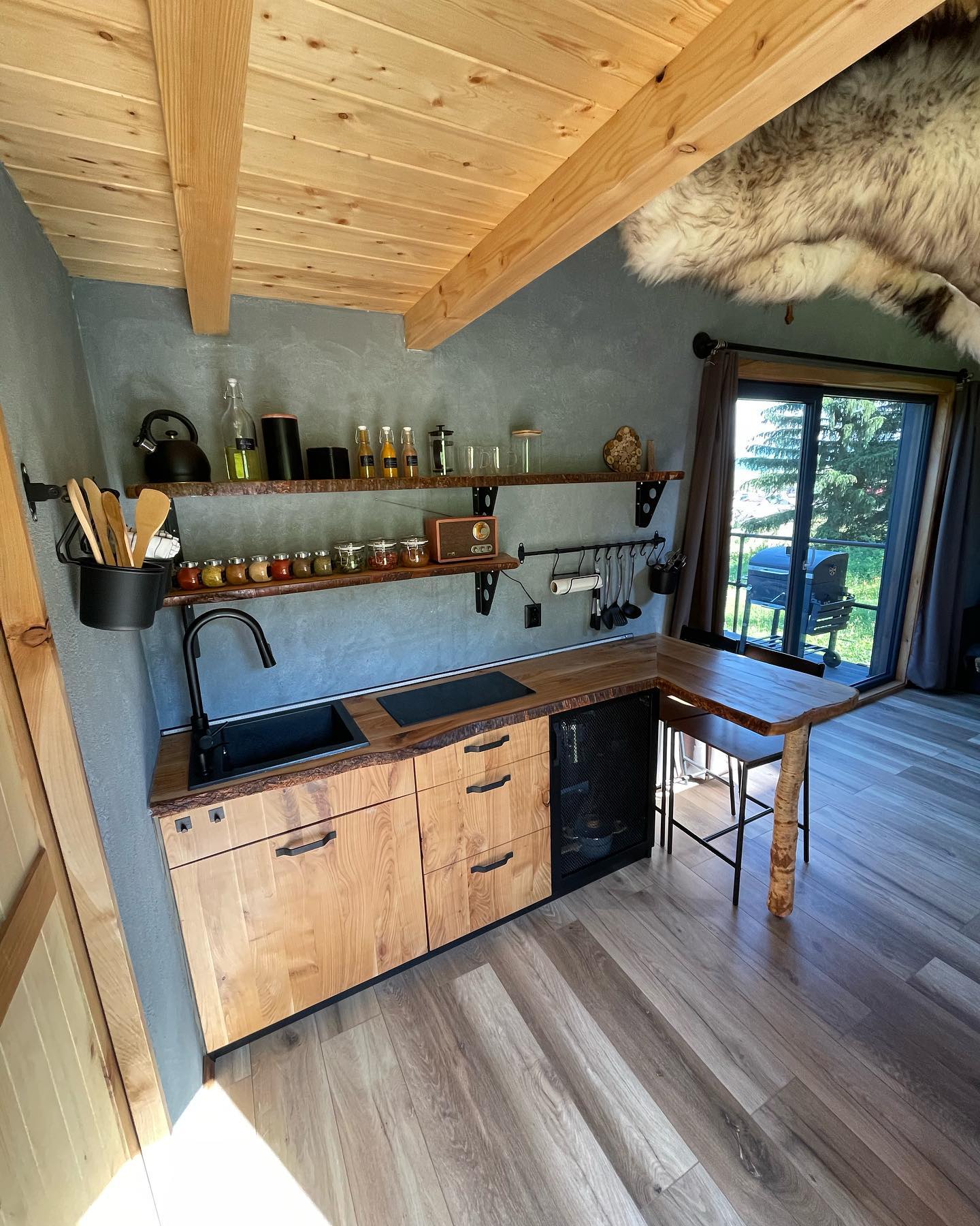 Rustic cabin tiny house kitchen with live edge wood countertops, matte black fixtures, plywood ceiling with exposed beams, a shelf of amber spice jars, and a vintage radio
