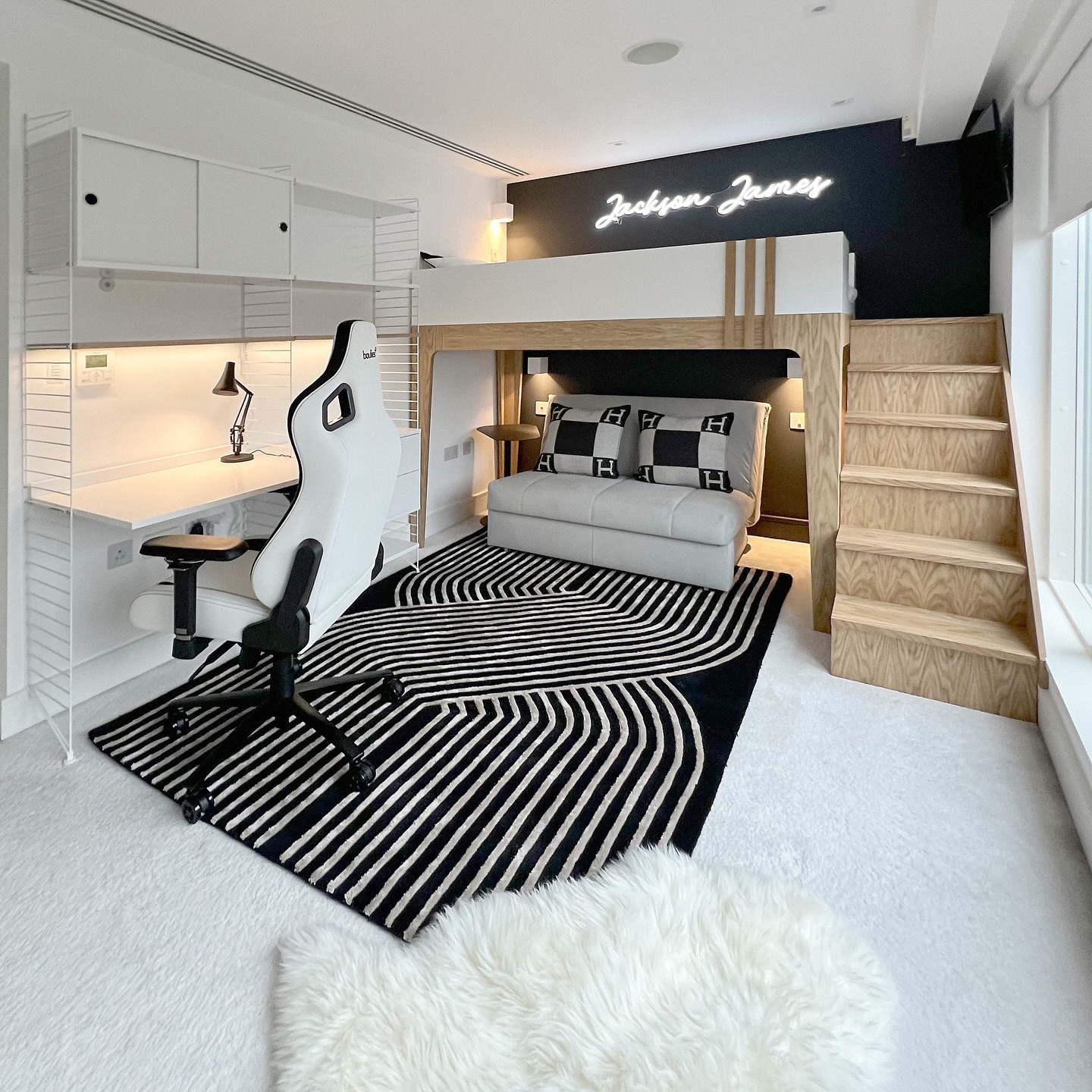 Alt: White lofted bed with wood staircase above a gray tufted sofa and black feature wall with a Jackson James neon sign, white floating desk and white racing-style gaming chair in the corner to the left