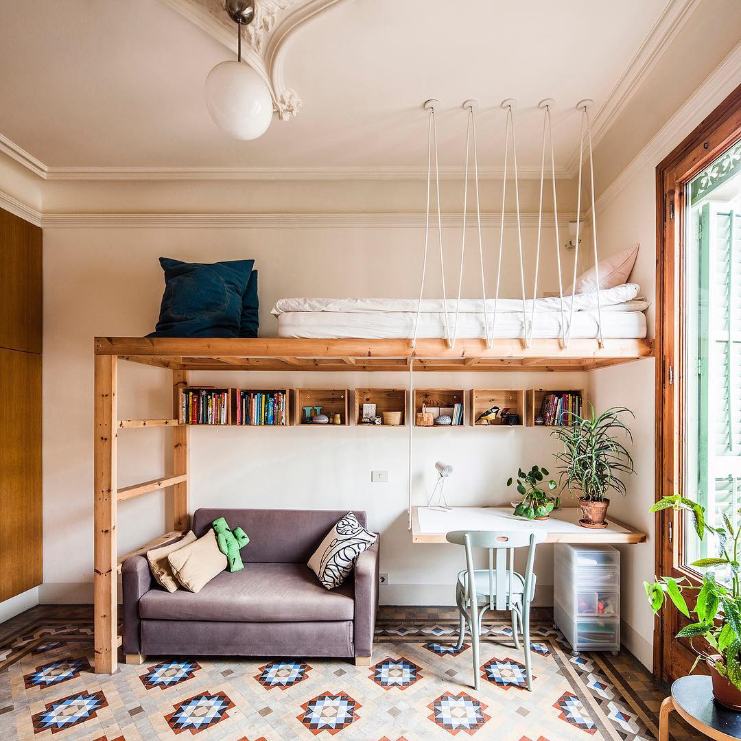 Alt: Wooden loft bed suspended from the ceiling with rope cables above a wall of pine book crates and a small white fold-down desk in a Barcelona apartment with ornate crown moulding and patterned tile floor