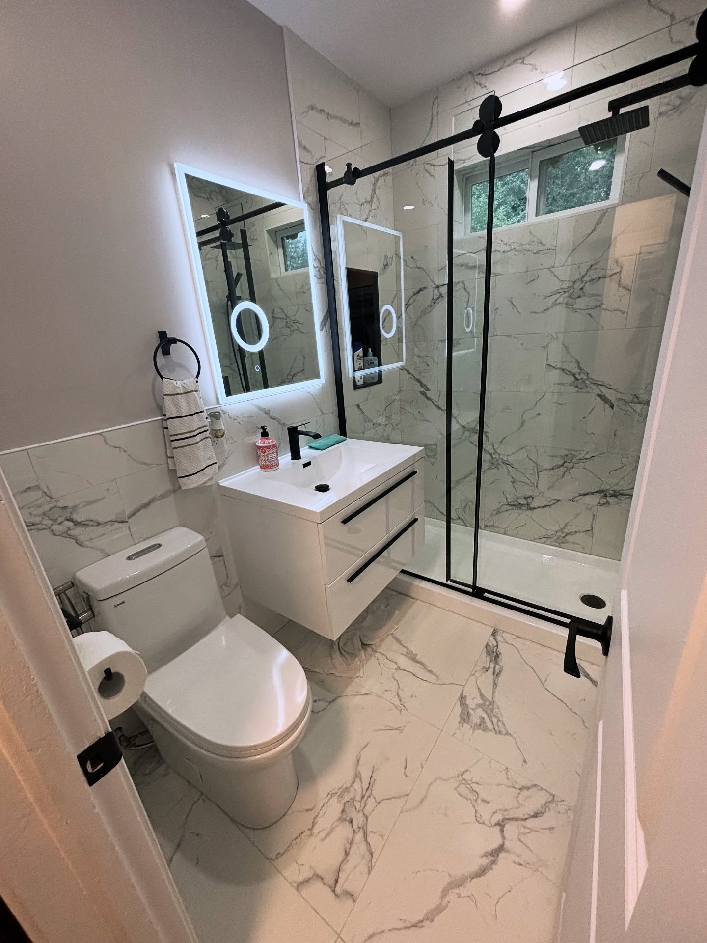 Small white marble tile bathroom with a black-framed sliding glass shower door, floating white vanity with black bar pulls, square LED backlit mirror, wall-hung toilet and black single-lever fixtures