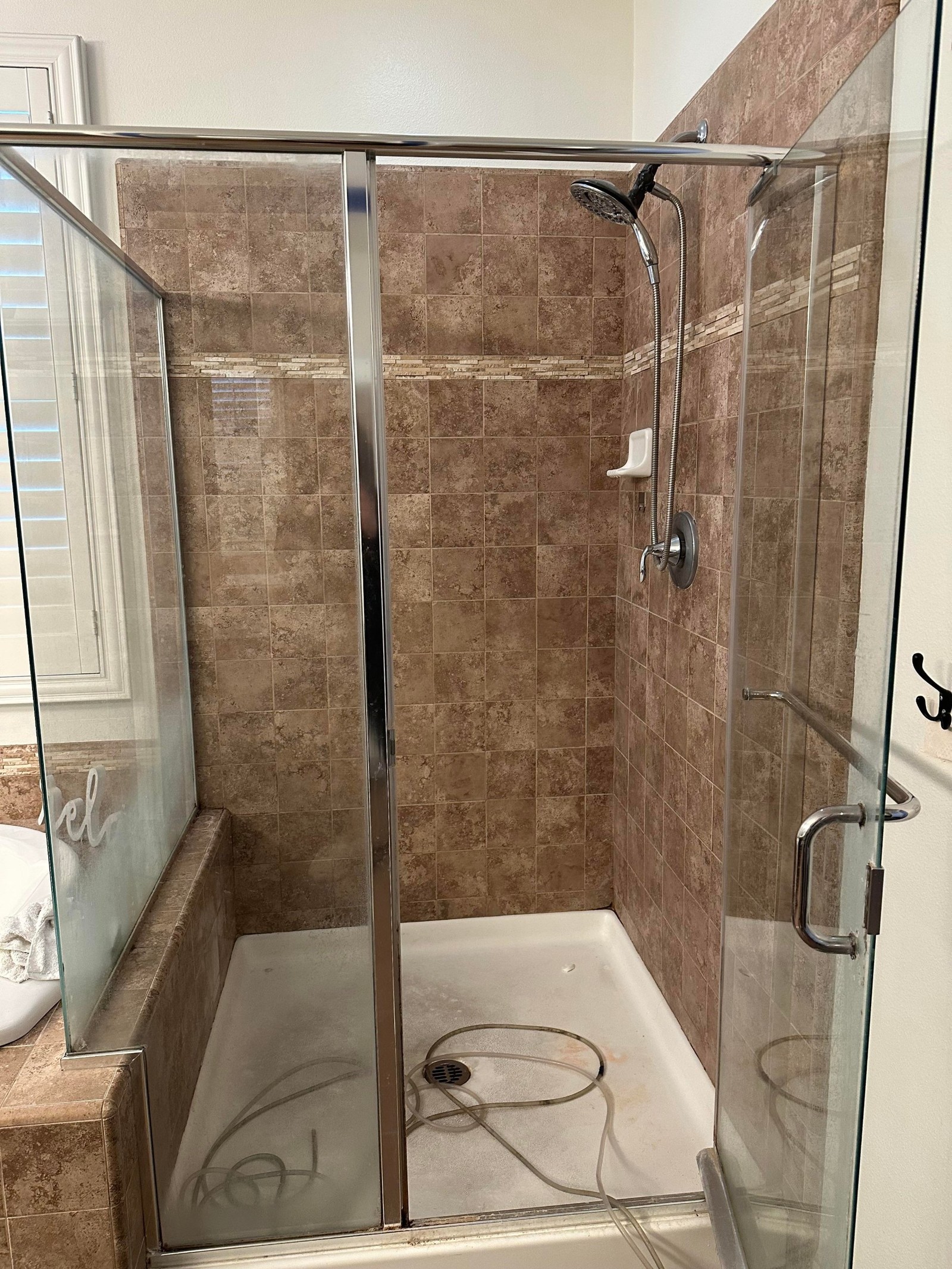 Shower stall with brown stone-look square tile on all three walls, a thin mosaic accent band running across, a chrome-framed glass door, a white plastic caddy on the back wall, and a white shower pan with coiled hoses on the floor.