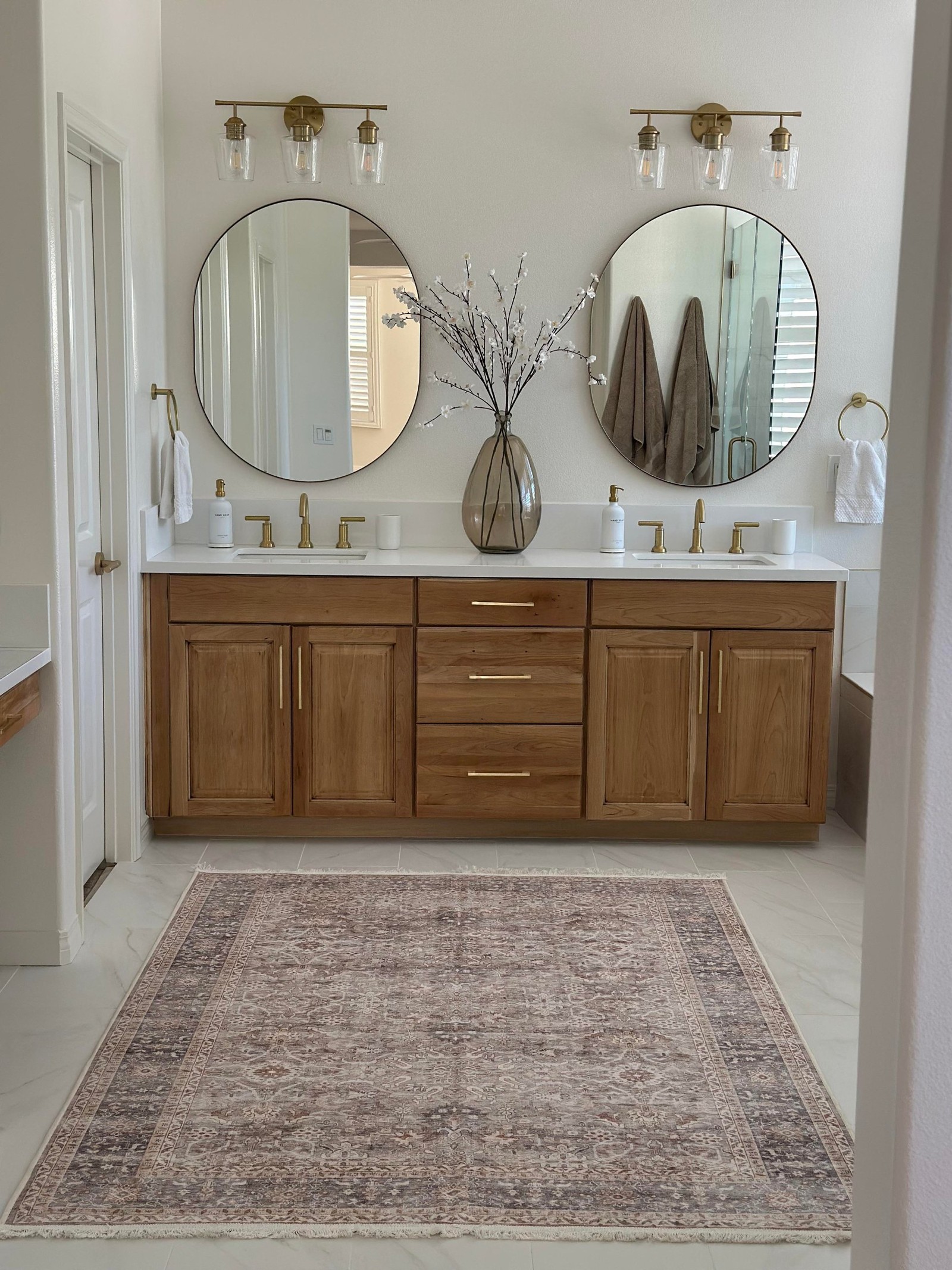 Head-on view of a double vanity with warm honey wood cabinet doors and drawers, long brass bar pulls, a white quartz counter with two undermount sinks, two round black-framed mirrors, brass sconces with clear glass shades, and white blossom branches in a smoky glass vase.