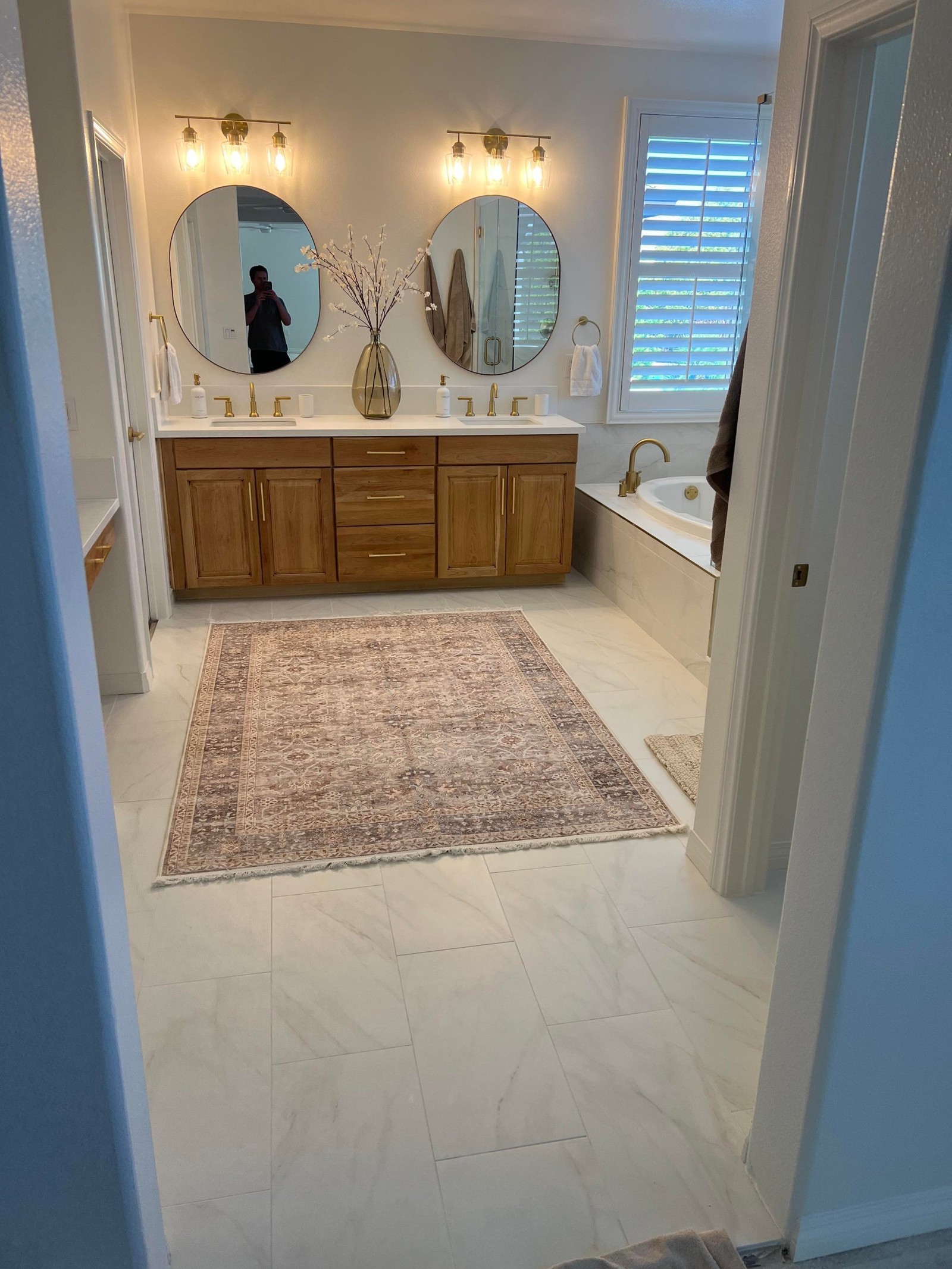 View from the doorway showing a patterned area rug in muted brown and cream on white marble-look floor tile, the honey wood double vanity with brass sconces and round mirrors, and the oval drop-in tub to the right.