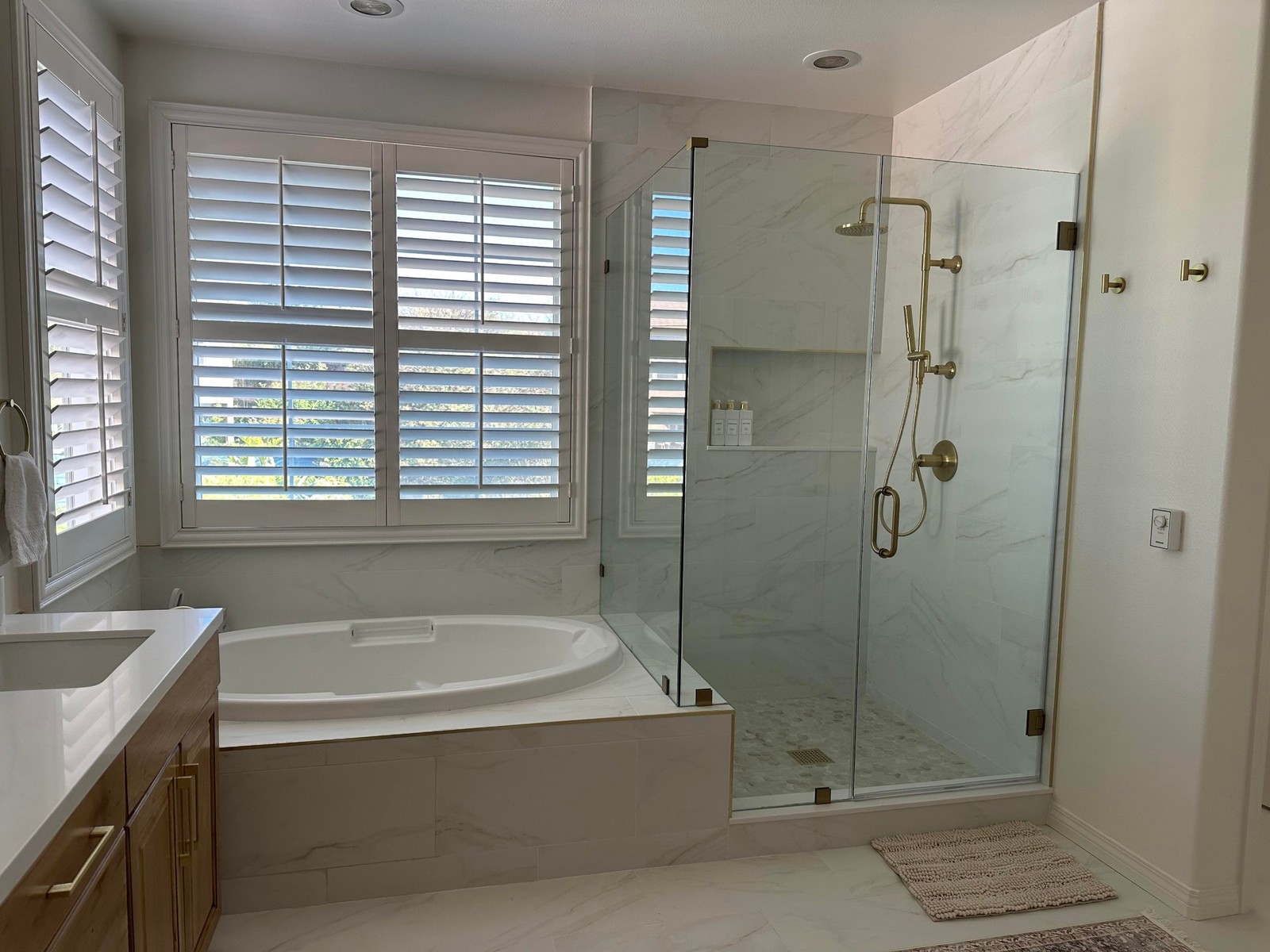 Finished bathroom with white marble-look tile on the walls and floor, an oval drop-in tub set into a white tile surround under the window, a brass tub filler, a frameless glass shower with brass hardware, white shutters, and warm honey wood cabinets on the left.