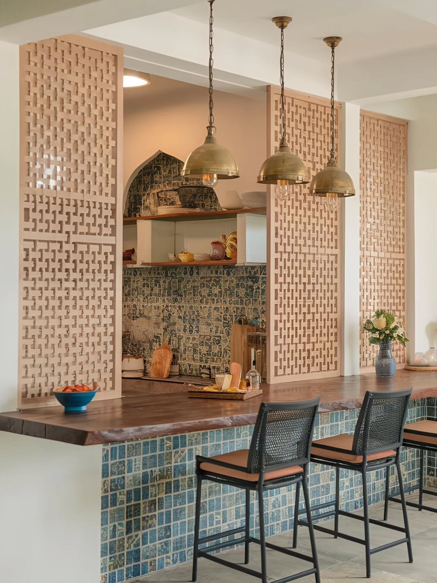 Open Mediterranean kitchen with carved pale pink stone screen partitions in a geometric basket-weave pattern, blue mosaic tile wrapping the bar front, three brass dome pendant lights on chains, a live-edge wood bar top, black rattan-back stools with rust cushions, and an arched mosaic tile niche in the background