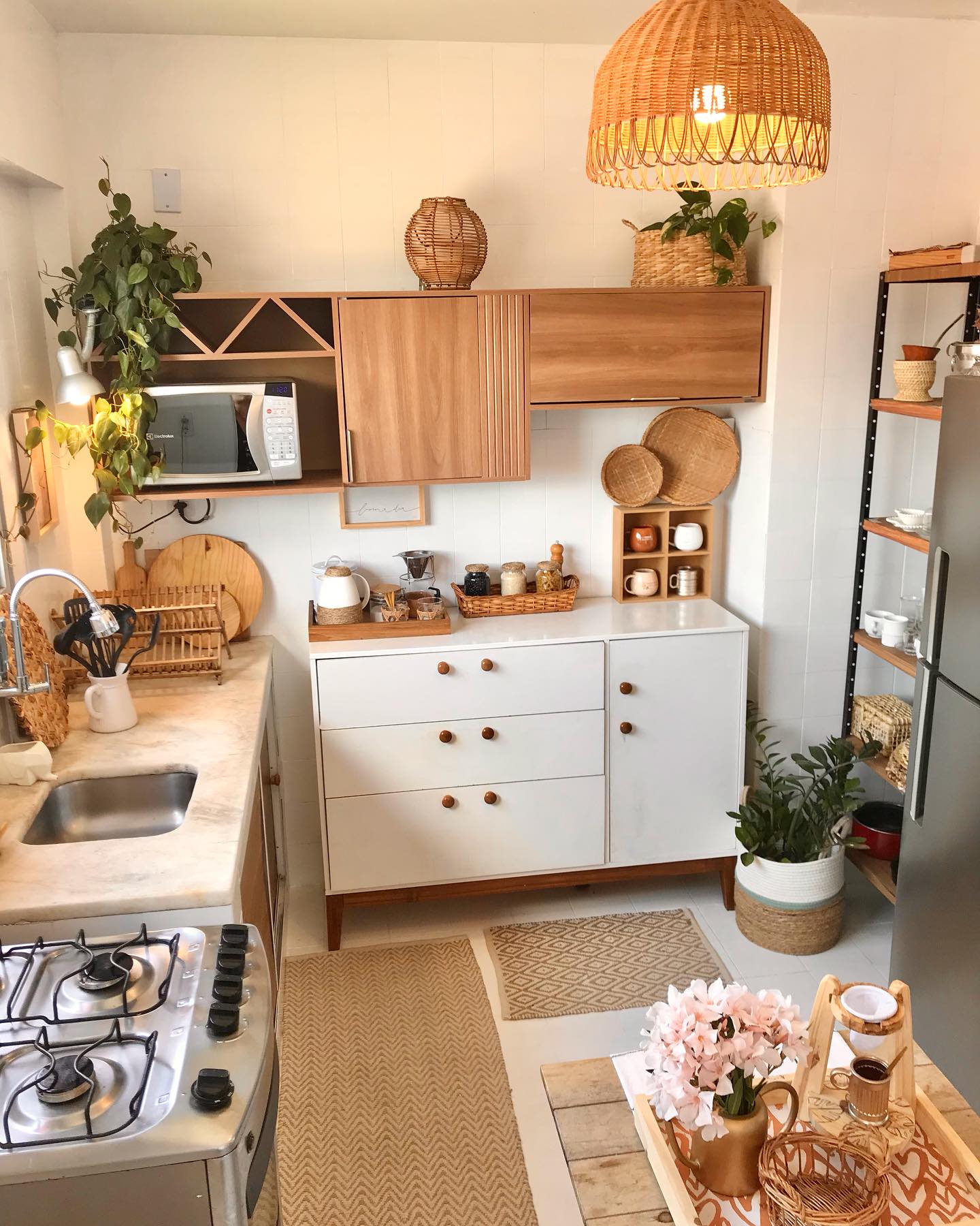 Small galley kitchen with mid-century slatted wood wall cabinet, large wicker dome pendant, trailing pothos plants, marble counter, white tile floor, jute runners, and a vintage microwave in the wall unit