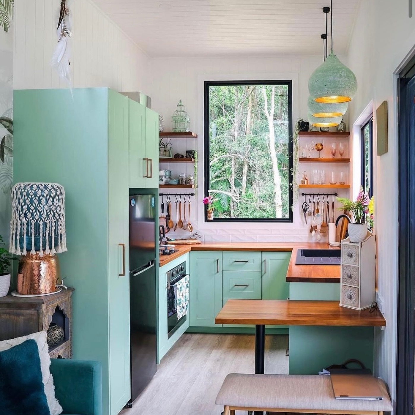 Mint green tiny house kitchen with butcher block counters, three woven pendant lights, open shelves with copper and glass accents, utensils hanging on wall hooks, and a large black framed window