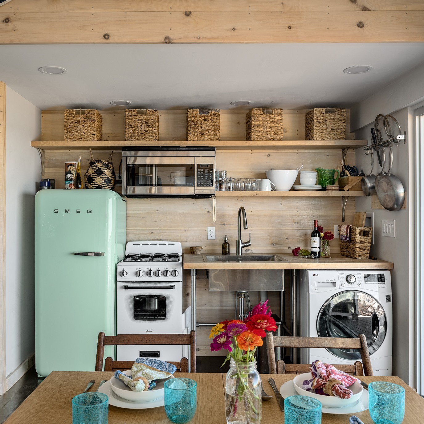 Tiny house kitchen with a pastel mint SMEG retro fridge, white Avanti gas range, open shelves topped with five woven baskets, stainless farmhouse sink, and a front-load LG washer tucked under the counter