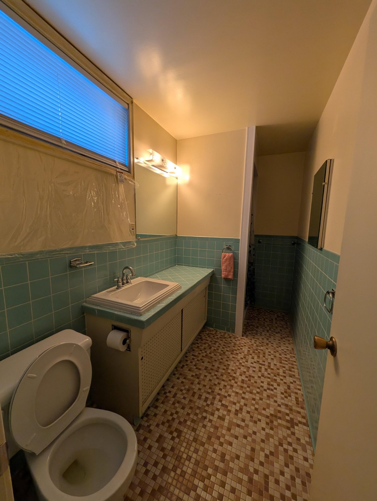 Dated bathroom with mint-green 4x4 wall tile, matching tiled countertop, pink-and-brown mosaic floor, a brass door knob, and a cheap vanity bar light.