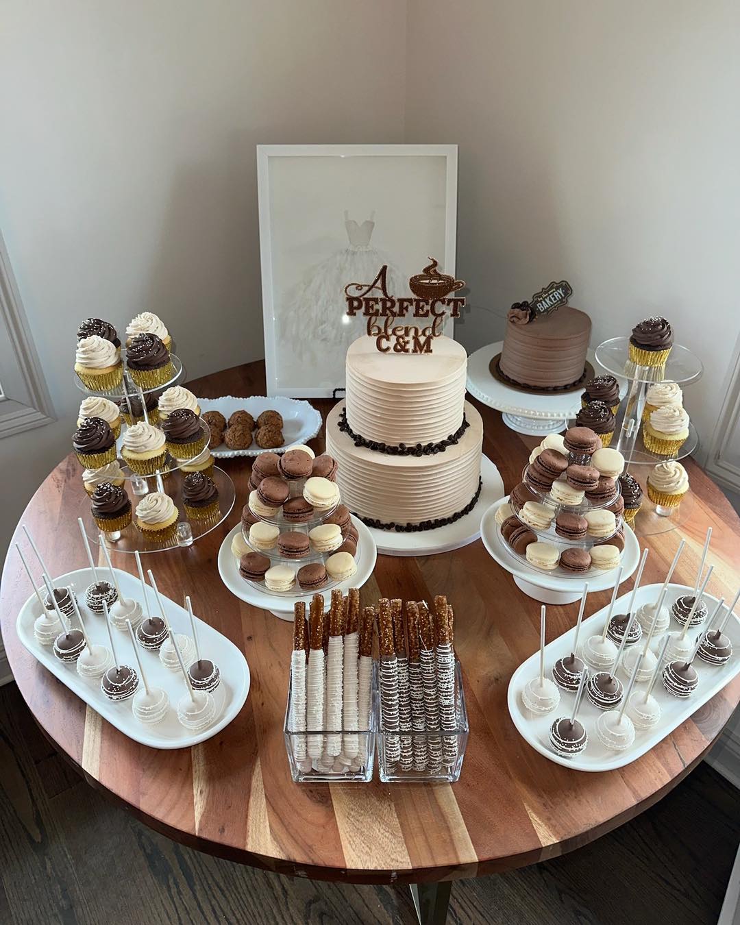 Mocha and neutral dessert table with two-tier cream cake, cream and chocolate macarons, cupcakes with chocolate frosting, chocolate dipped pretzel rods, and cake pops on a wood round table