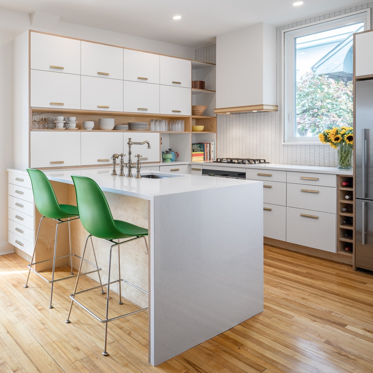 Modern white flat front kitchen with birch plywood edges exposed, long brass bar pulls, open shelving with white dishware, small white square mosaic tile backsplash, bright green molded shell counter stools with chrome legs, and warm maple hardwood floor