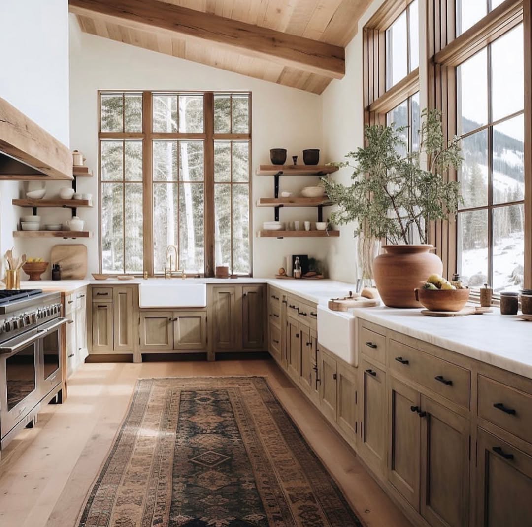 Mountain cabin kitchen with light wood cabinets, two farmhouse sinks, large wood-framed windows looking out at snow and evergreens, open floating shelves on black iron brackets, and a vintage dark red runner on wide-plank oak floors