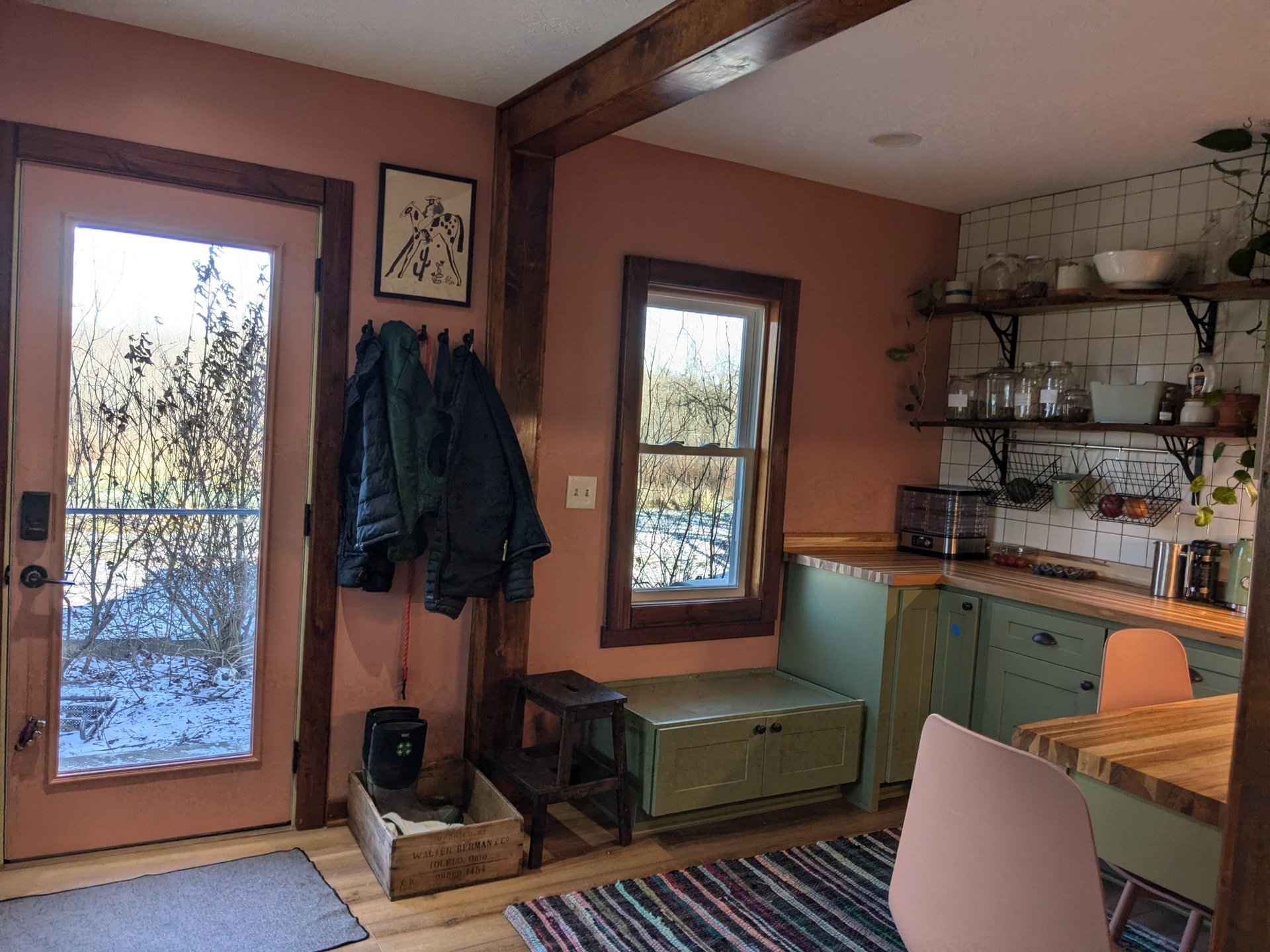 Finished mudroom corner with coral pink walls, a built-in sage green storage bench under a window, coat hooks with jackets, a vintage wooden crate, and open shelving in the adjoining baking nook.
