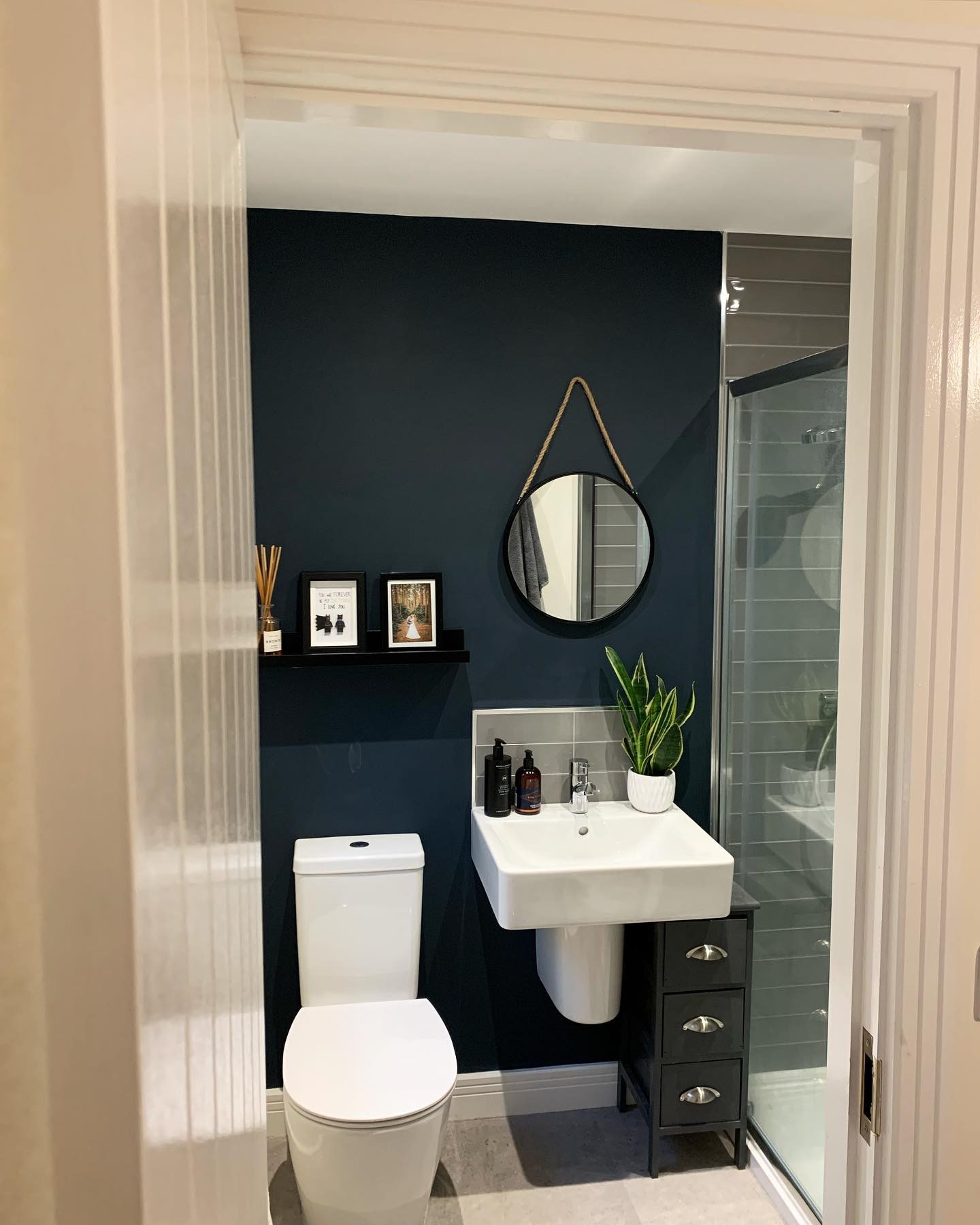 mall bathroom with a single navy blue accent wall behind the toilet, round rope-hung black mirror, black floating shelf with framed prints, white wall-mounted sink, and a snake plant on the sink