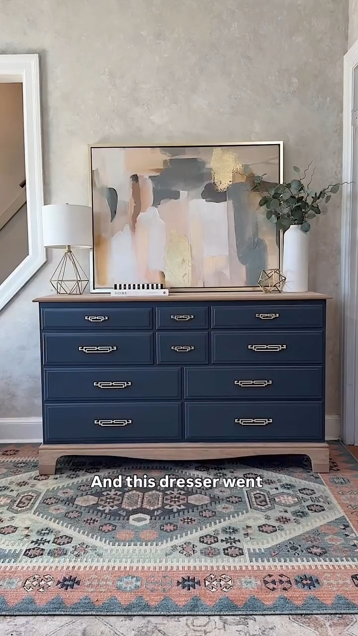 A deep navy dresser with a natural wood top and slim silver bar pulls, styled against a greige wall with a large abstract painting in navy, cream, pink and gold tones, a geometric wire lamp with a white shade, a white vase of eucalyptus branches, a small HOME sign, and a Southwestern kilim-style rug on the floor