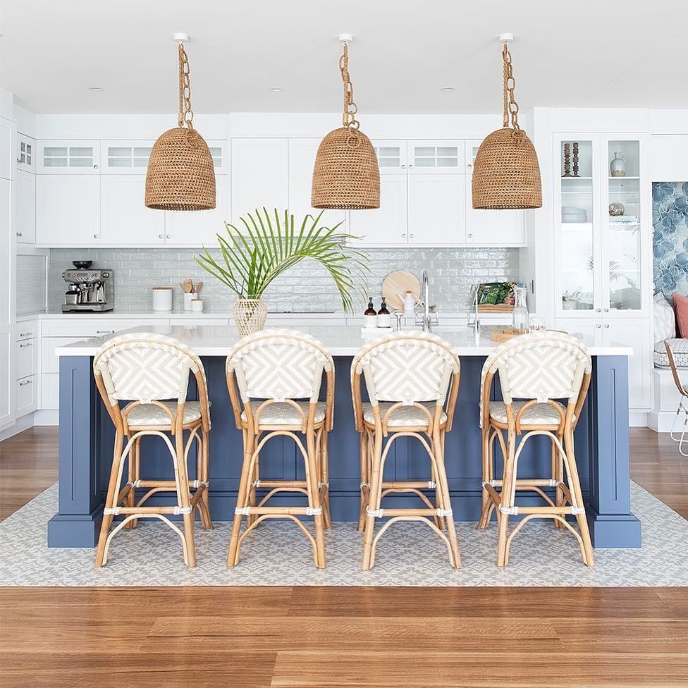 White coastal kitchen with a deep navy painted island, four rattan French bistro counter stools with woven backs and seats, three large woven basket pendants, pale textured subway tile backsplash, and warm wood flooring