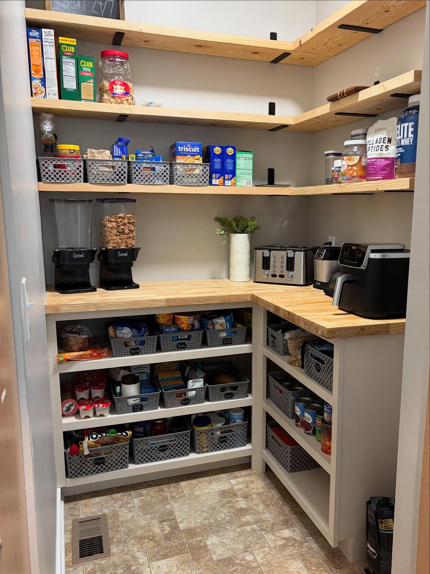 Walk-in pantry with a tall white shelving tower full of navy and white striped fabric bins, L-shaped acacia butcher block counters lined with small appliances, clear labeled canisters, and an angled wire can rack on the right wall