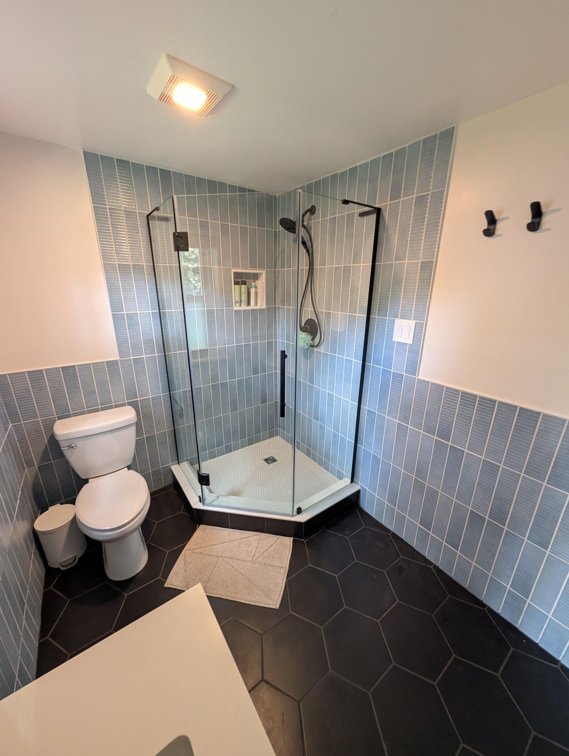 Frameless neo-angle glass shower set in the corner with dusty blue vertical stacked tile covering the walls up to the ceiling, a built-in niche holding toiletries, a white penny tile shower pan, black matte shower fixtures, and large black hexagon tile on the bathroom floor.