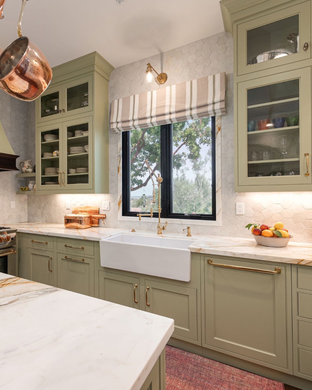 Olive sage green kitchen with brass gooseneck faucet, white farmhouse apron sink, striped Roman shade, marble counters with gold veining, and a hanging copper pot
