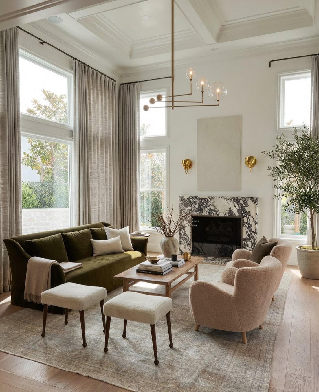 Elegant modern farmhouse living room with olive green velvet sofa, blush boucle swivel chairs, black-veined marble fireplace, brass pendant chandelier, and tall linen drapes