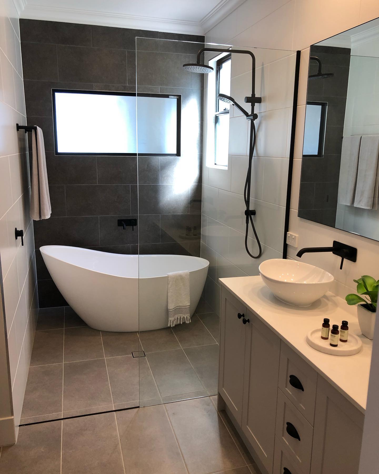 Small open wet room style bathroom with a white freestanding slipper tub against a dark grey feature tile wall, walk-in shower with black rain head, white shaker vanity with a round white vessel bowl sink and a single frameless glass panel divider