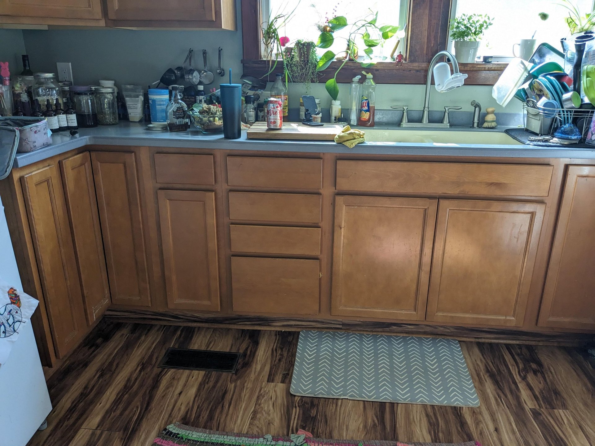 Wide view of the before kitchen with orange-stained oak cabinets, grey laminate countertops, a cluttered sink wall packed with jars and plants, and dark laminate wood-look flooring.